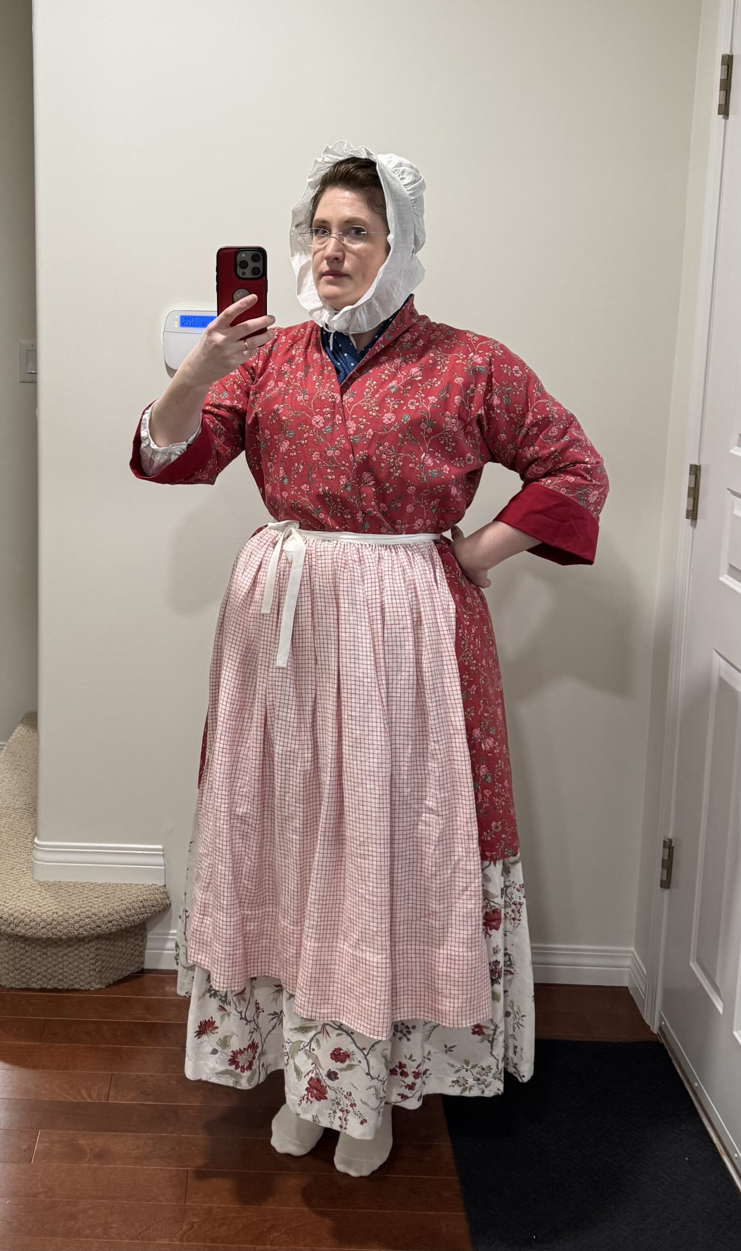 A woman faces the camera wearing a tomato red chinz bedgown, a red and white check apron and a cream chintz petticoat. She is also wearing a lappet cap on her head.