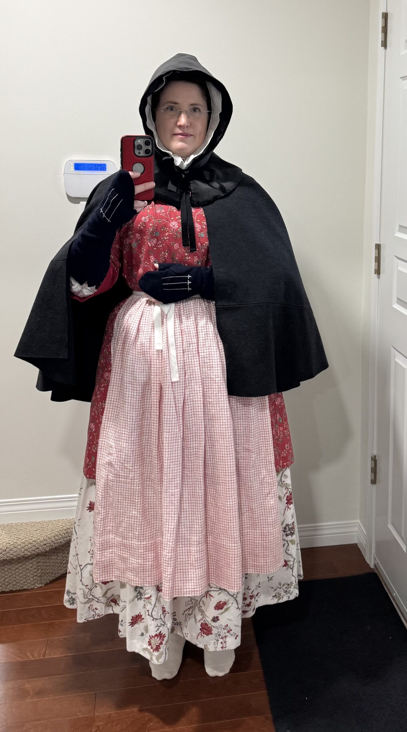 A woman faces the camera wearing a tomato red chinz bedgown, a red and white check apron and a cream chintz petticoat below a grey wool short cloak and blue wool mitts.
