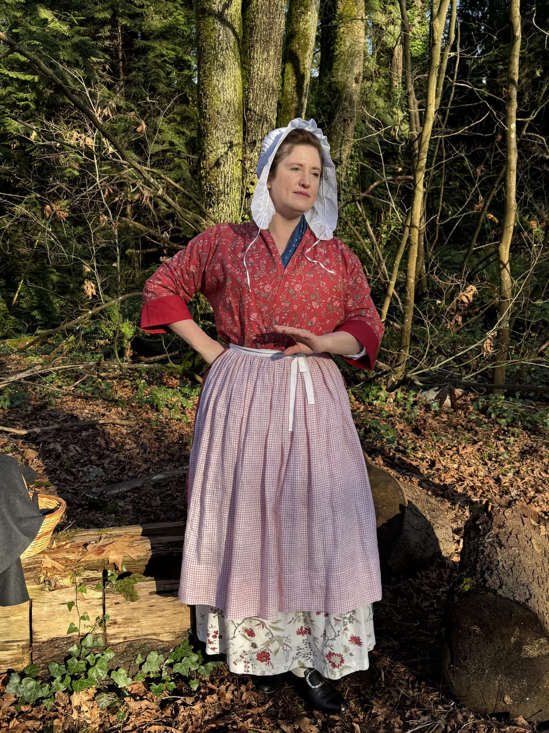 A woman stands in a grove of trees, wearing a red chintz bedgown, and a plaid petticoat. On her head is a white linen lappet cap with the strings untied.