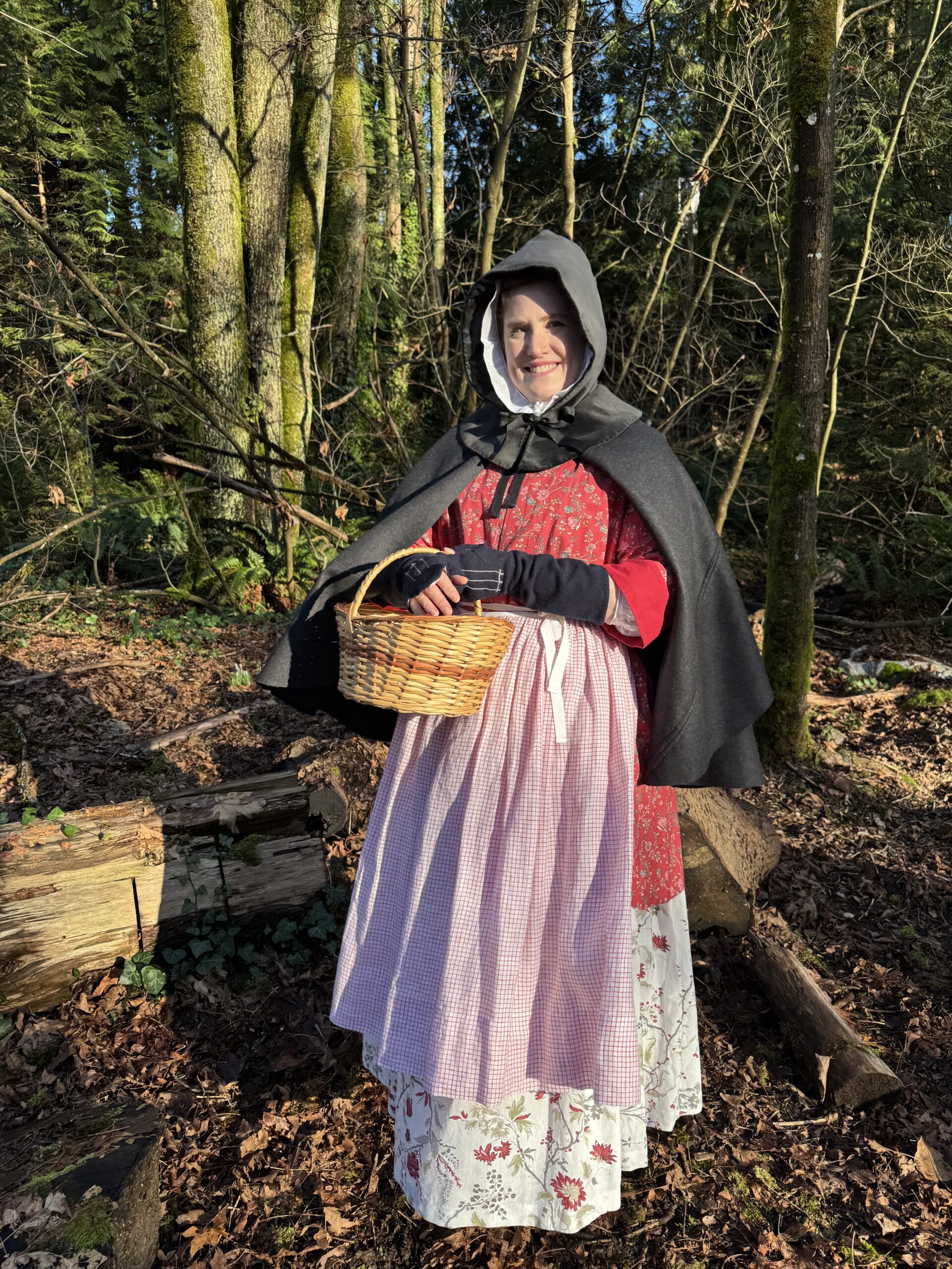 Tabubilgirl stands facing the camera. She wears a 1770s chintz ensemble formed of bedgown and petticoat, and a red check apron, grey wool short cloak and blue wool mitts. In her arms she carries a woven wicker basket.