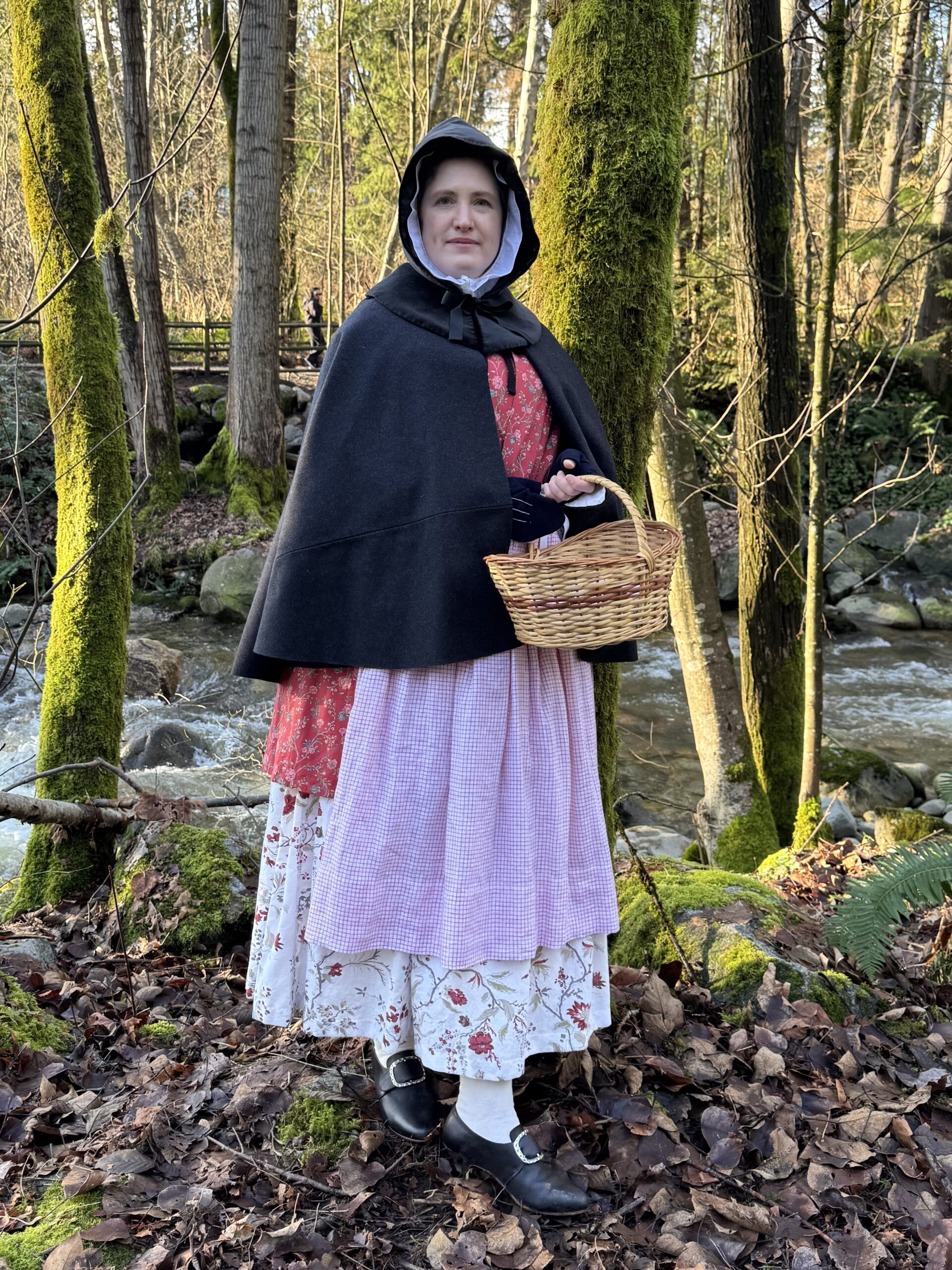 A woman stands facing the camera wearing a 1770s winter-weather chintz ensemble - petticoat, bedgown, apron, short cloak and velvet hood.