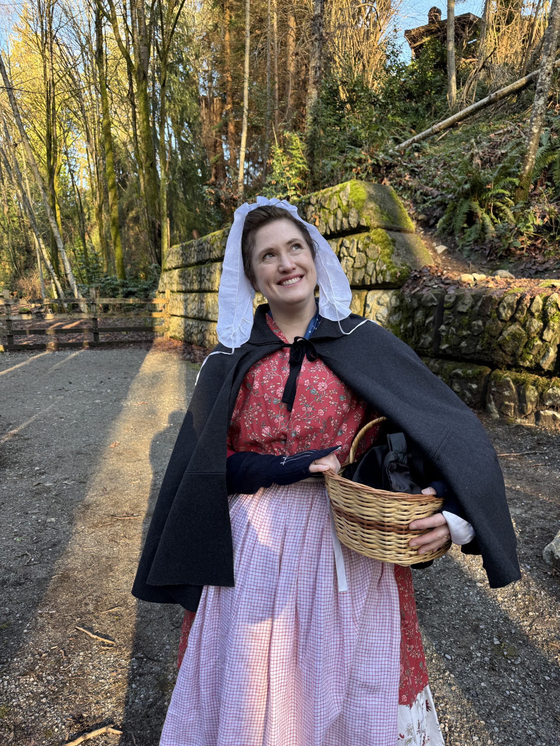Tabubilgirl looks away to the right. she is wearing a red chintz bedgown and red check petticoat below a short grey wool cloak, and she carries a basket under her arm.