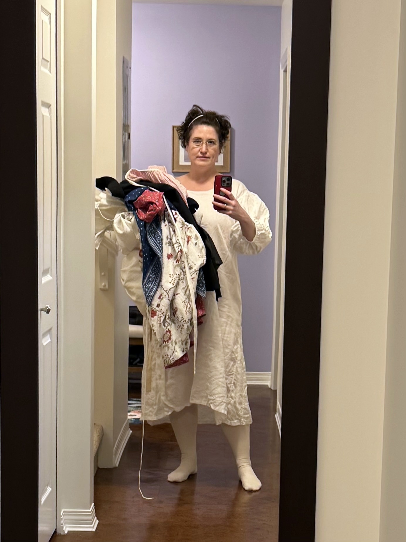 Tabubilgirl is taking a selfie in a mirror. She is wearing a mid-18th century shift and carrying a stack of chintz garments.
