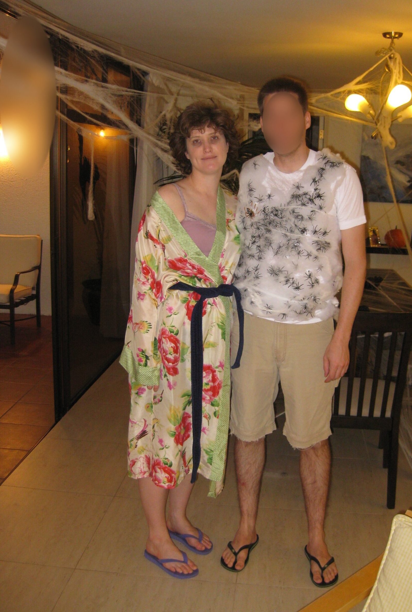 A woman wearing a bathrobe and slippers stands next to a man wrapped in white fiberfill and black plastic spiders. They are clearly at a halloween party.